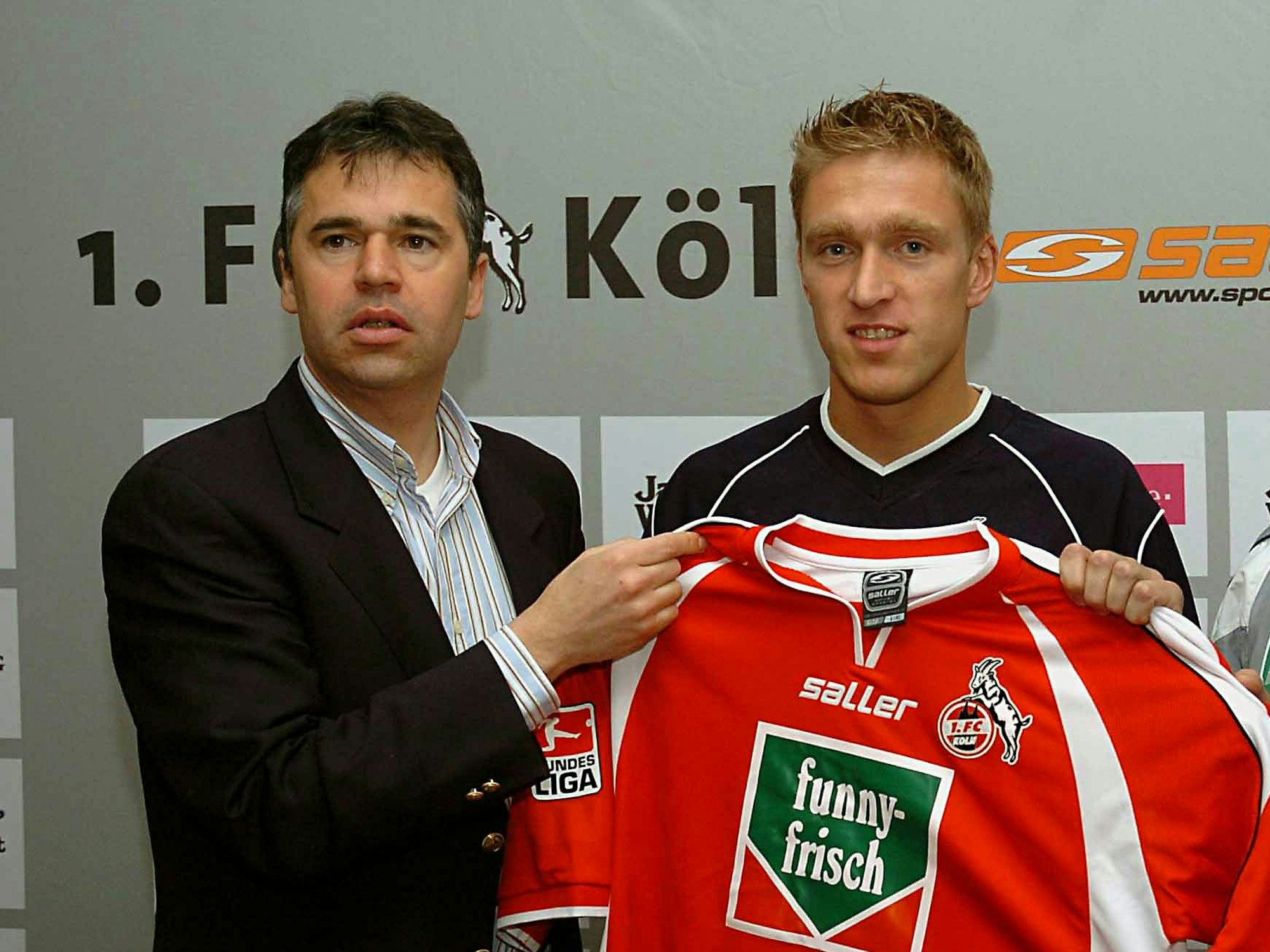 Andreas Rettig steht neben Christian Rahn. Sie halten ein FC-Trikot hoch.