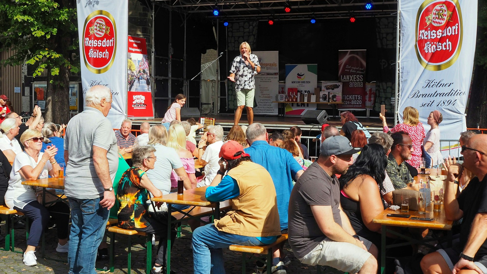 Menschen sitzen an Bierbänken vor der Bühne, auf der ein Mann singt.