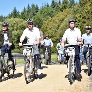 Die Politiker fahren mit dem Rad über einen Weg. Im Hintergrund sind Bäume zu erkennen.