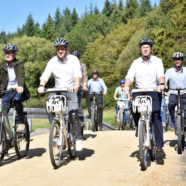 Die Politiker fahren mit dem Rad über einen Weg. Im Hintergrund sind Bäume zu erkennen.