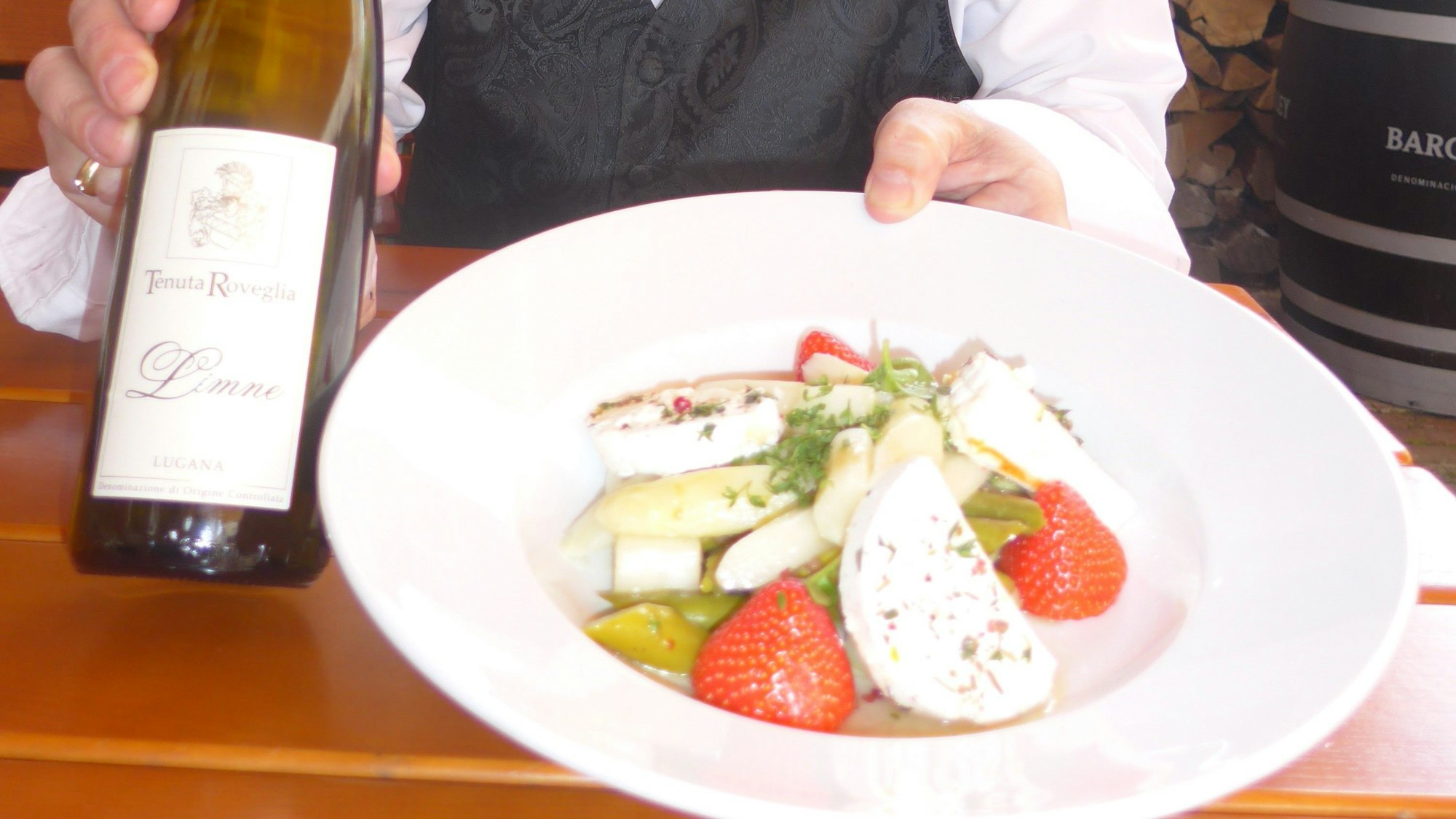 Ein Mann hält einen Teller mit Essen und eine Flasche Wein in die Kamera.