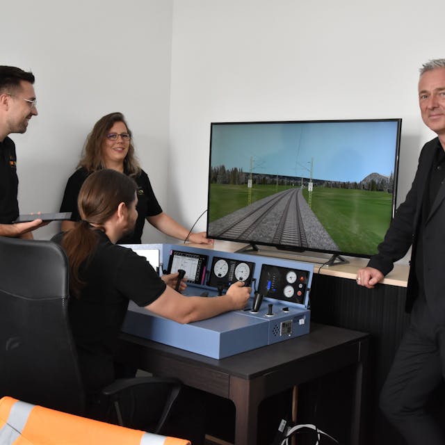 Vier Personen stehen am Simulator eines Lok-Führerstandes. Einer von ihnen steuert die Lok über eine Strecke, die auf dem Bildschirm zu sehen ist.