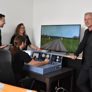 Vier Personen stehen am Simulator eines Lok-Führerstandes. Einer von ihnen steuert die Lok über eine Strecke, die auf dem Bildschirm zu sehen ist.