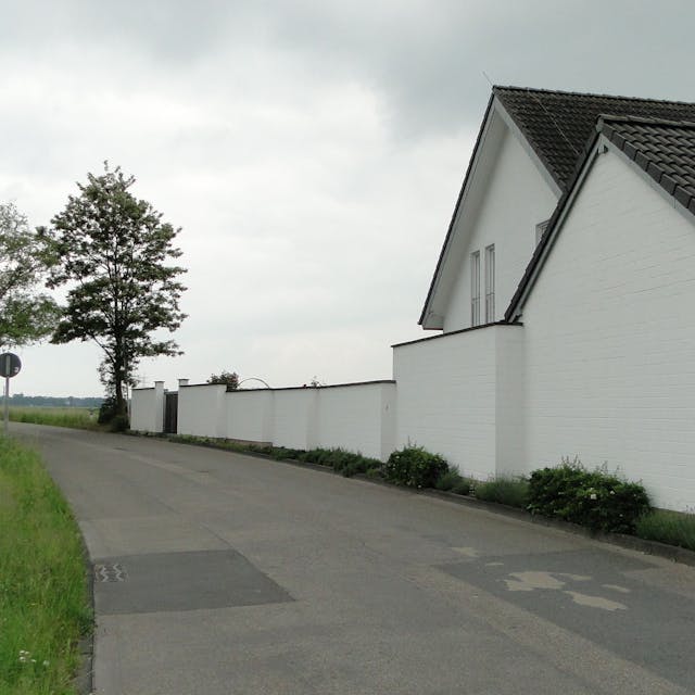 An einer Landstraße liegt ein weißes Haus mit Mauer und auf der anderen Seite eine Wiese mit vereinzeltem Baumbestand.