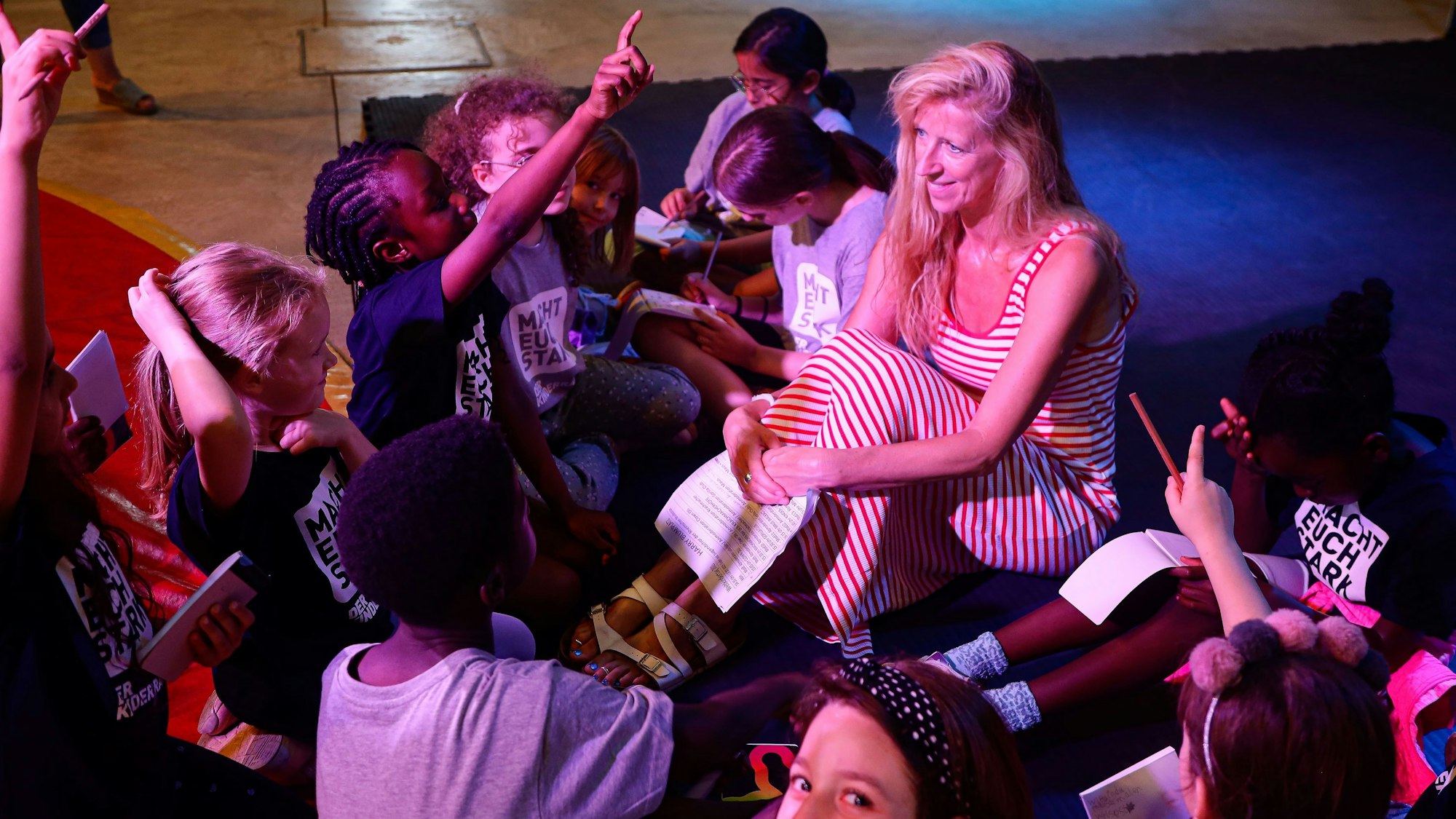 WDR-Moderatorin Insa Backe sitzt auf dem Boden, um sie herum haben sich die Jungen und Mädchen des „Linoclub“-Kinderrates versammelt. Sie proben gemeinsam ihren Auftritt beim Weltkindertag.