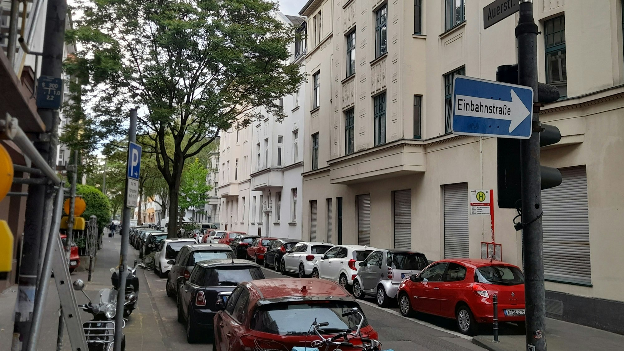 Schmale Einbahnstraße mit parkenden Autos auf beiden Straßenseiten und einem großen Baum auf der linken Straßenseite
