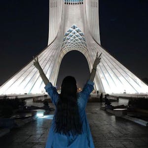 Eine junge Frau steht ohne das vorgeschriebene Kopftuch vor dem Azadi-Turm (Freiheitsturm) und zeigt mit beiden Händen das Sieges-Zeichen.