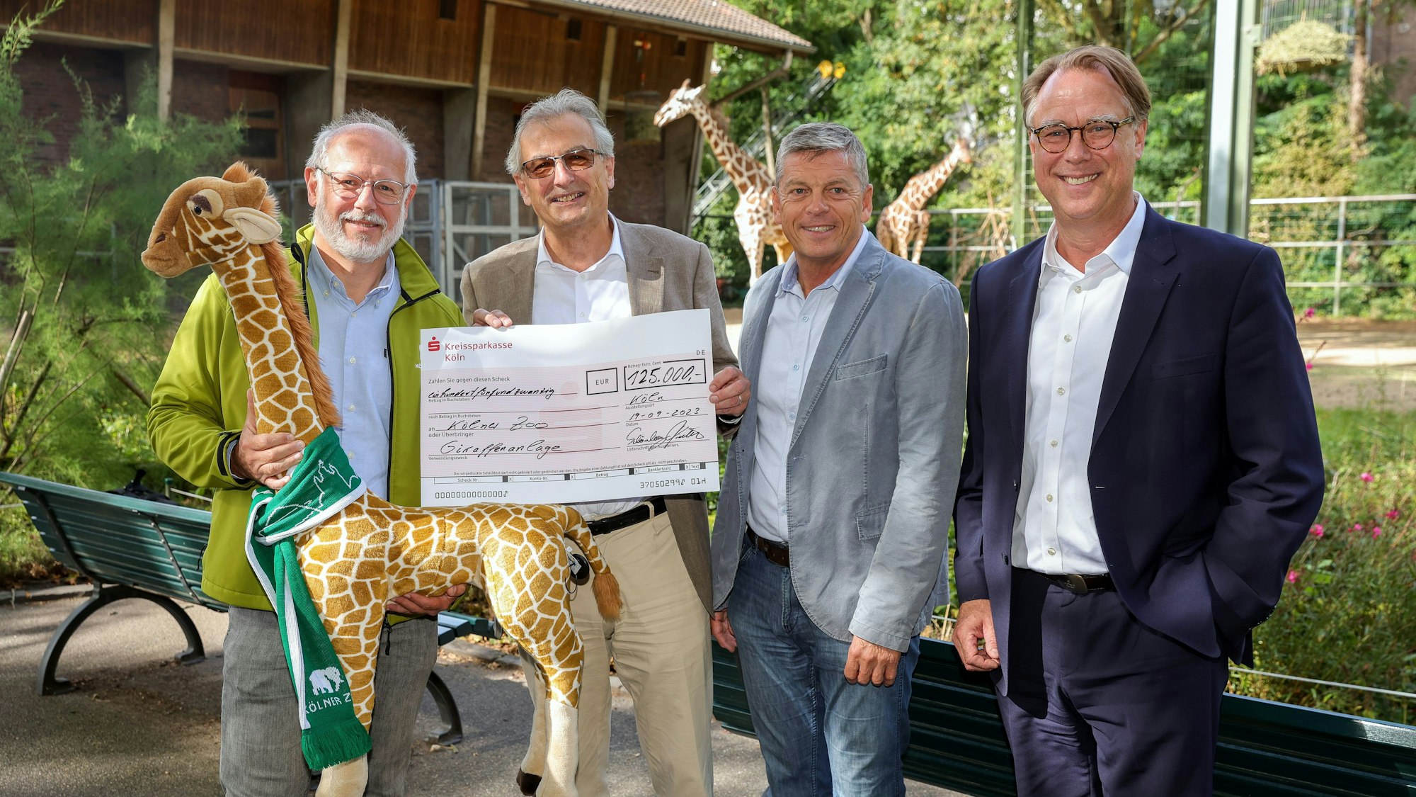 Vier Männer zeigen einen Spendenscheck vor der großen Giraffenanlage im Kölner Zoo.