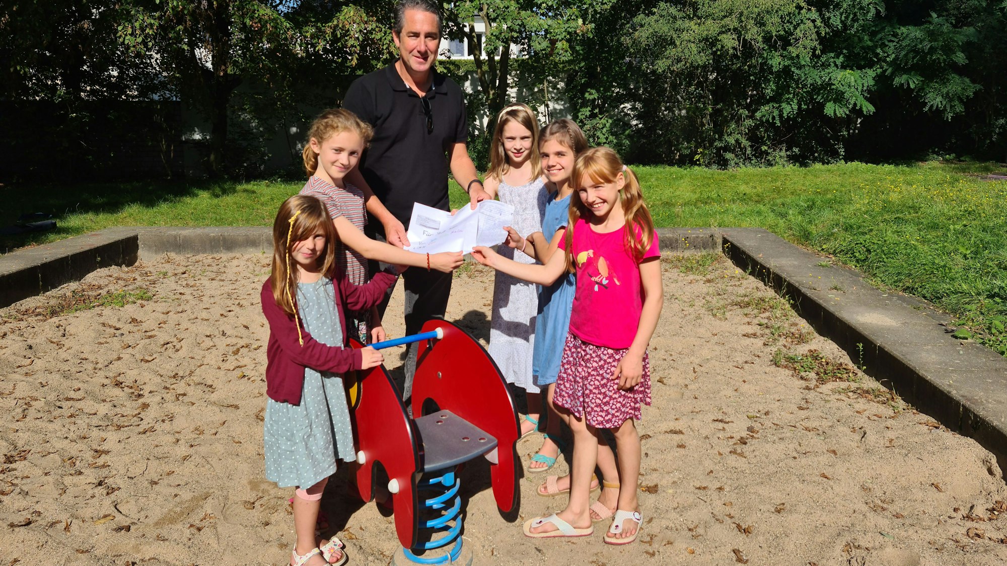 Fünf Mädchen stehen links und rechst von einem Wipptier in einem Sandkasten. Sie übergeben einem jungen Mann eine Liste mit Unterschriften.
