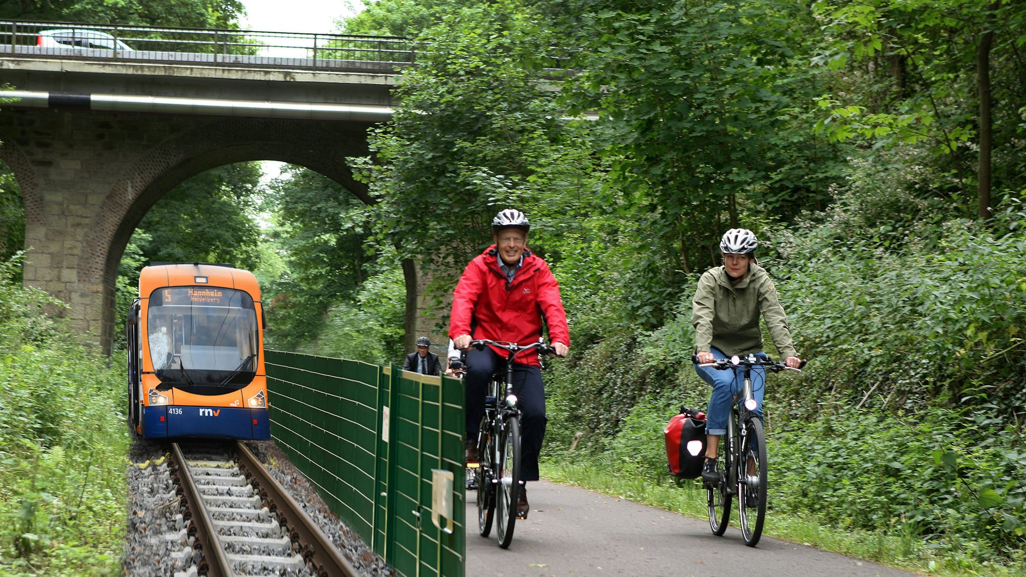 Zwei Fahrradfahrer fahren auf einem Rad-Gehweg auf einer ehemaligen Bahntrasse, daneben fährt – durch einen Zaun abgetrennt – eine Straßenbahn.