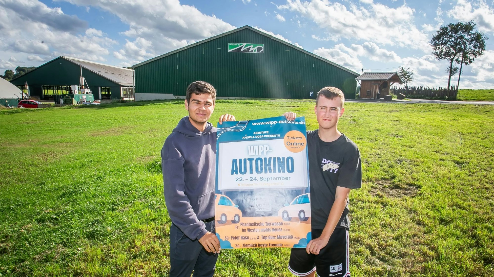 Zwei junge Männer stehen auf einer Wiese und halten ein Veranstaltungsplakat für das erste Wipperfürther Autokino in den Händen. Im Hintergrund ist eine große Scheune eines Landwirtschaftsbetriebs zu sehen.