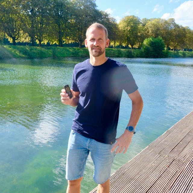 Ein Mann steht auf einem Steg an einem See, in der Hand hält er ein kleines Telefon.
