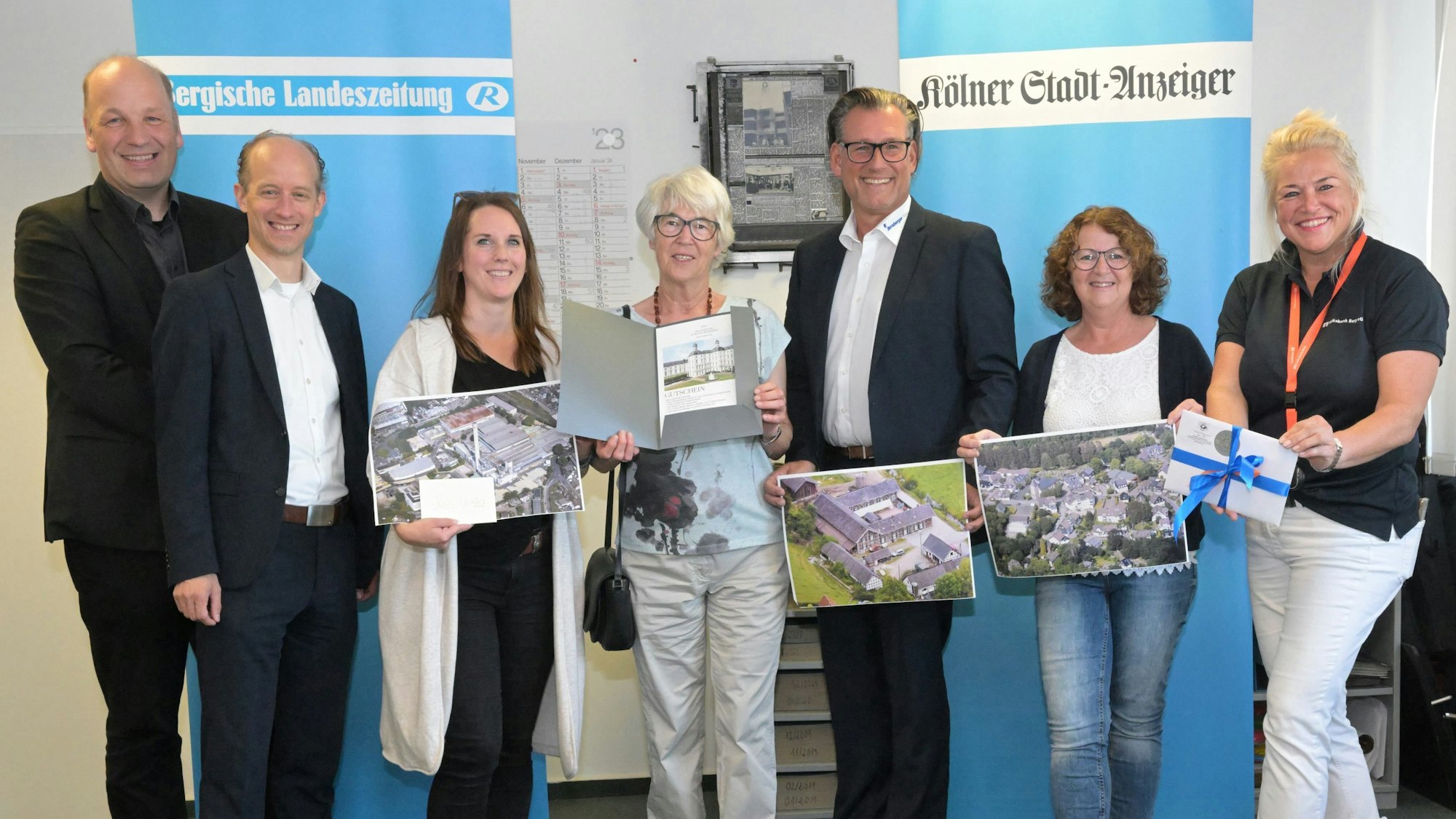 Menschen Stehen mit Luftbildern und Geschenken in den Händen vor Bannern von Bergischer Landeszeitung und Kölner Stadt-Anzeiger.
