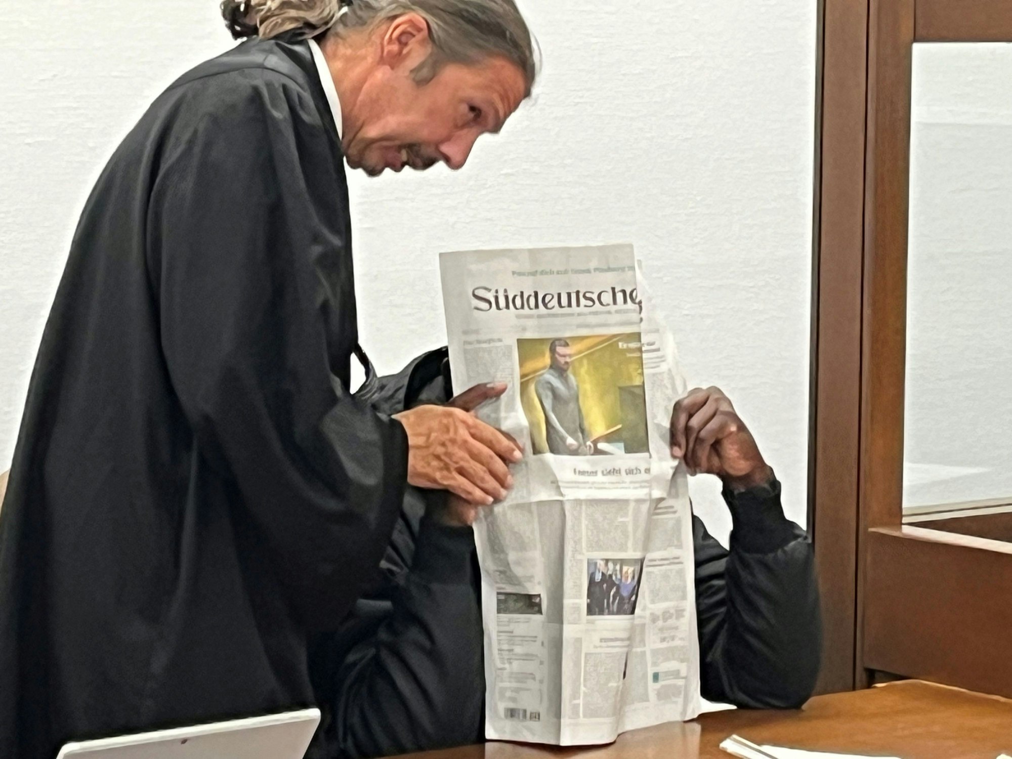 Der Beschuldigte hält sich eine Zeitung vor das Gesicht, neben ihm steht sein Anwalt.