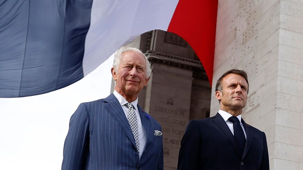 König Charles III (l) von Großbritannien und Emmanuel Macron, Präsident von Frankreich, stehen unter einer Frankreich-Flagge am Arc de Triomphe.
