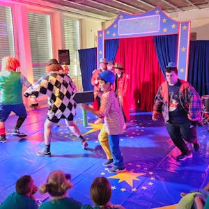Bunt kostümierte Erwachsene und Jugendliche laufen in einer kleinen Zirkusmanege im Kreis.