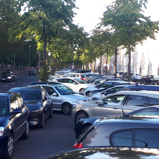 Auf der Mittelallee einer baumbestandenen Straße parken viele Autos.