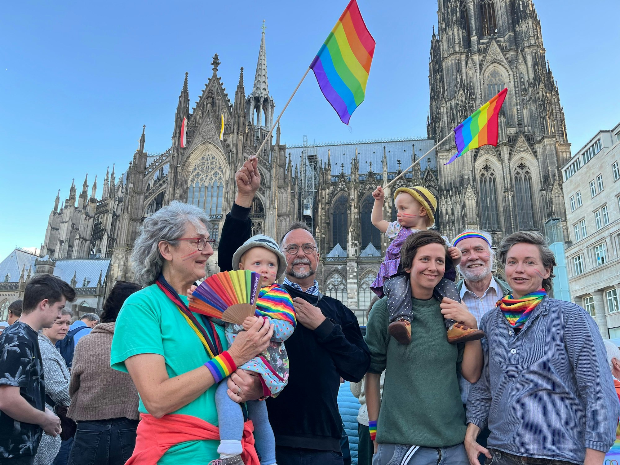 Zwei Frauen, drei Großeltern und zwei kleine Kinder mit Regenboggenflaggen vor dem Kölner Dom