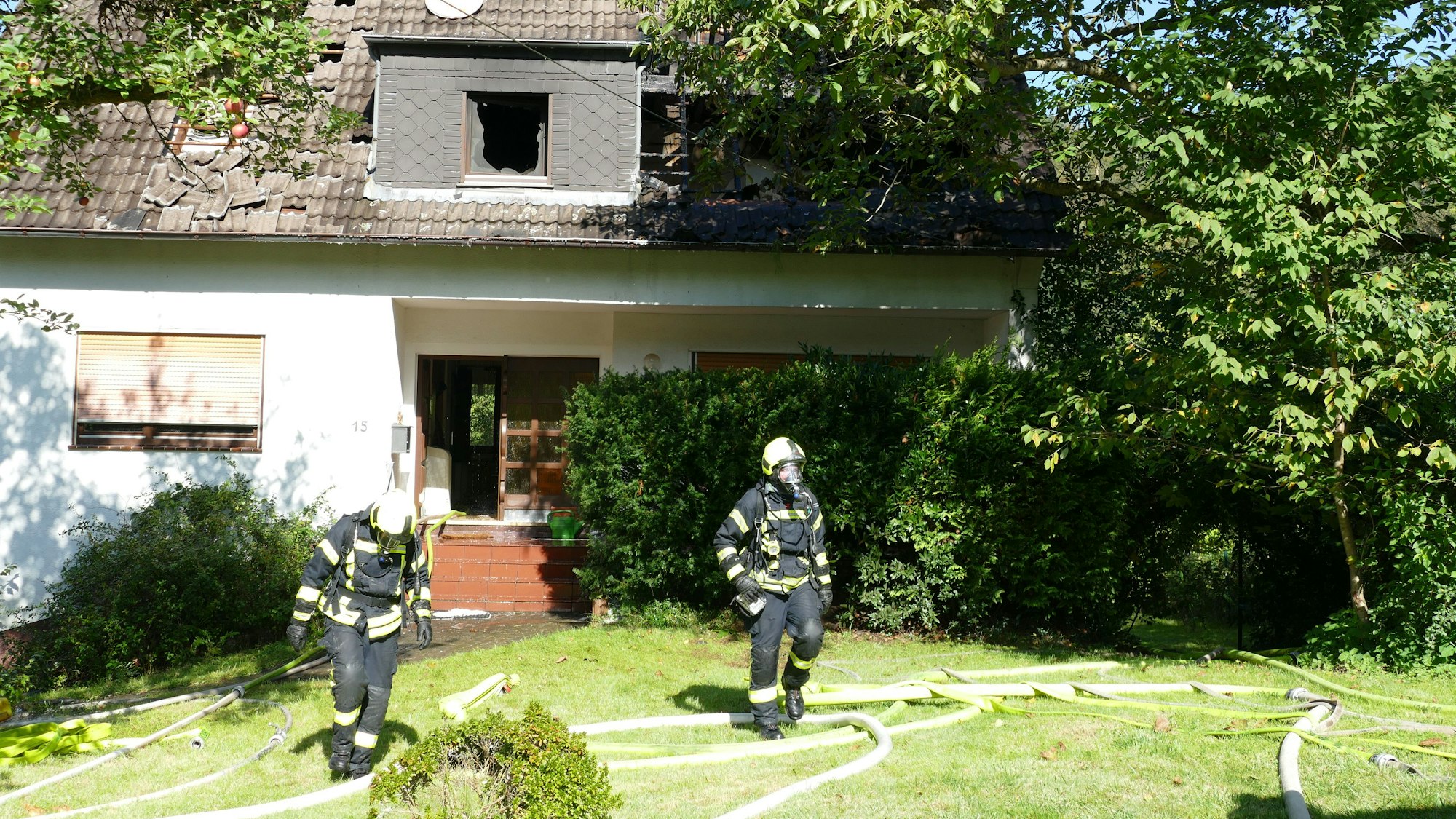 Zwei Feuerwehrleute in einem Garten, hinter ihnen ist ein Einfamilienhaus, in dessen Dach große Löcher sind, die Dachlatten sind verkohlt, die Fenster geborsten.
