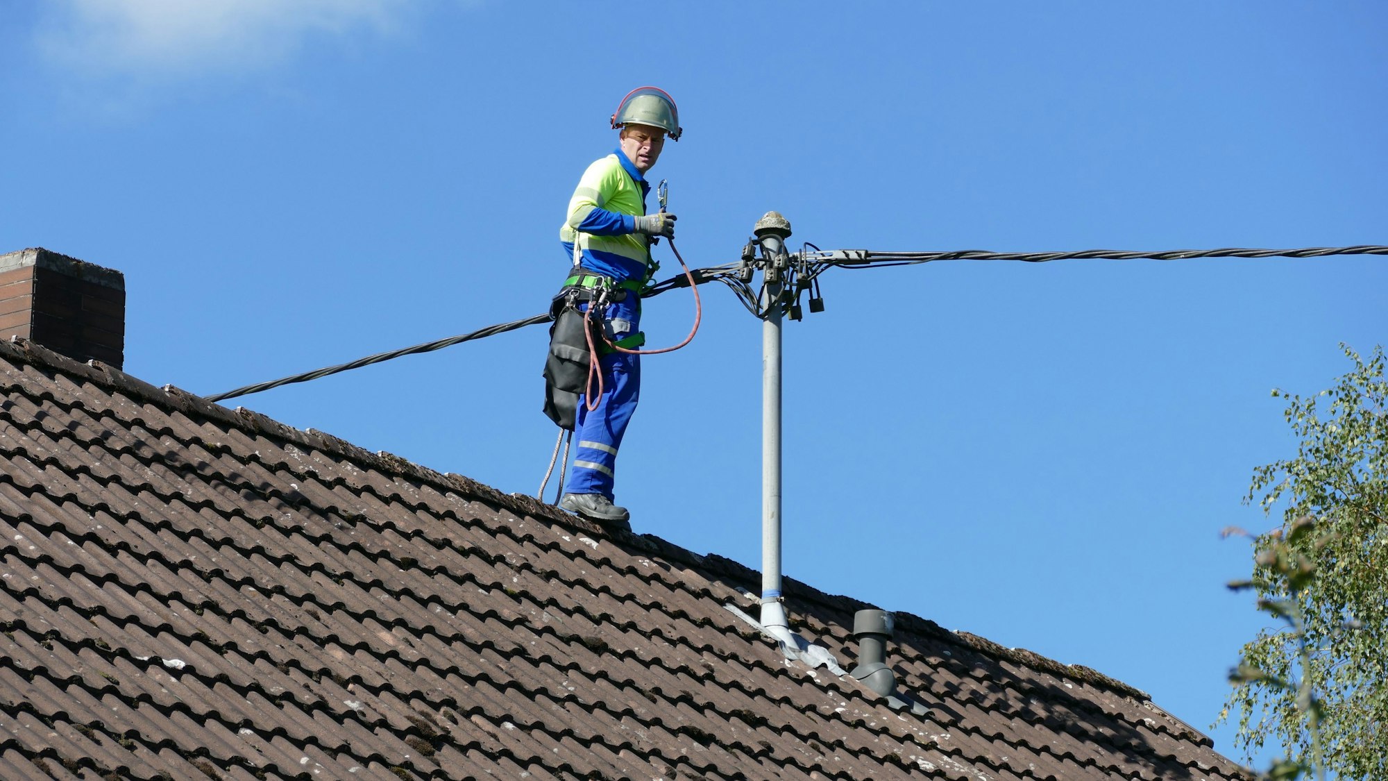 Ein Mann im blauen Overall und mit Helm steht auf dem Dachfirst an der Stromleitung.