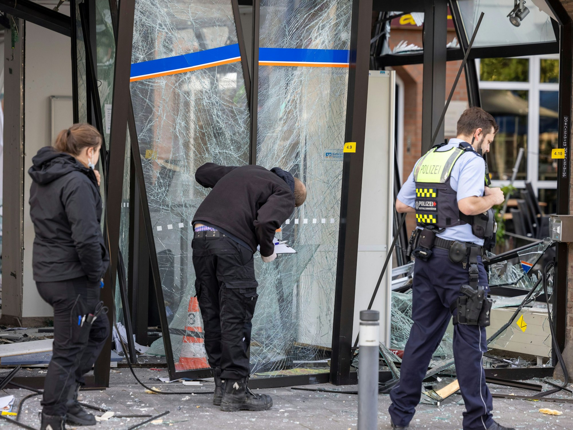 Drei Einsatzkräfte stehen vor der beschädigten Glasfassade der Bankfiliale, ein Beamter untersucht die geborstene Eingangstür.