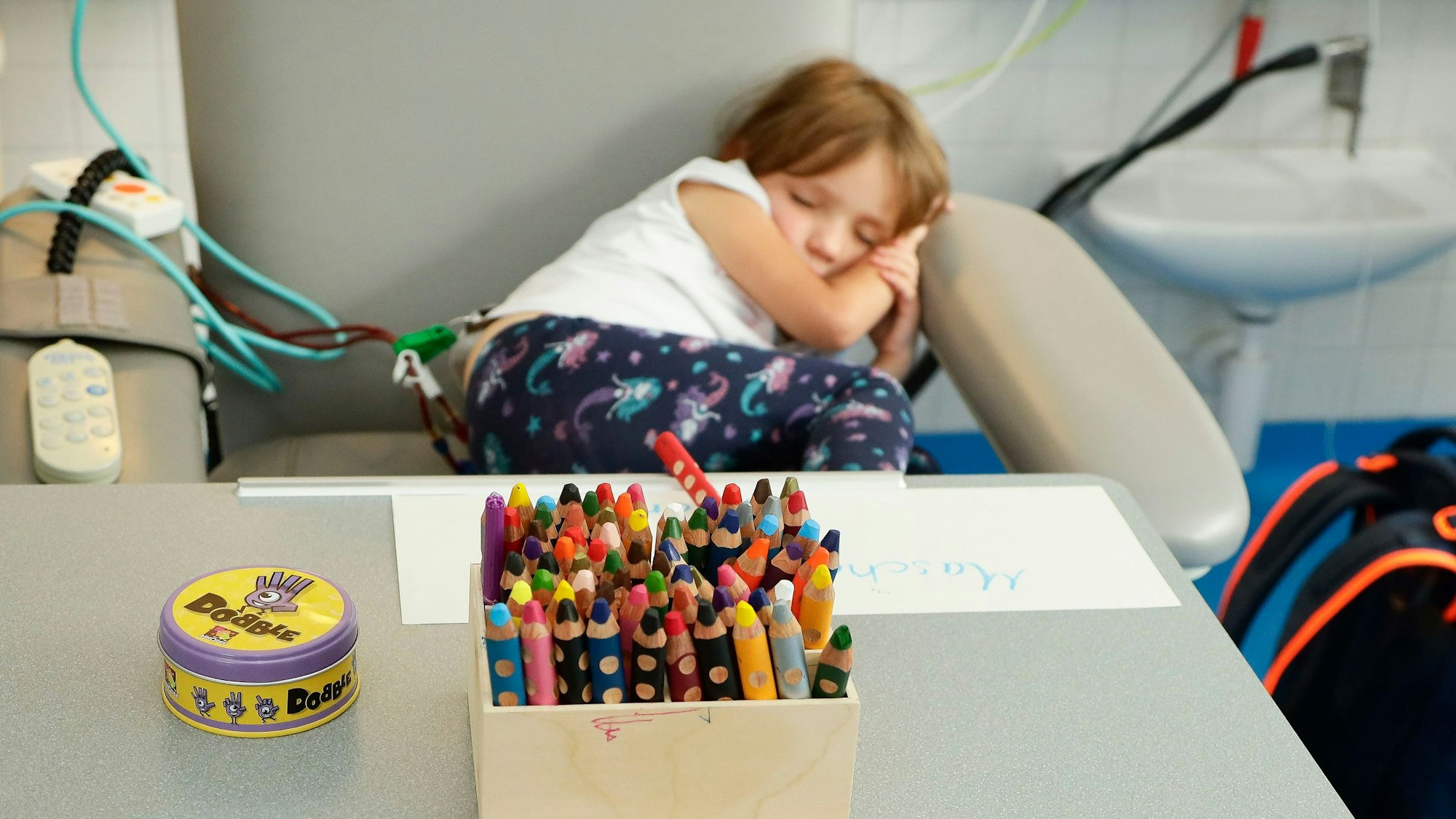 Ein kleines Mädchen liegt schlafend an der Dialyse, vor ihr steht eine Kiste voller Buntstifte