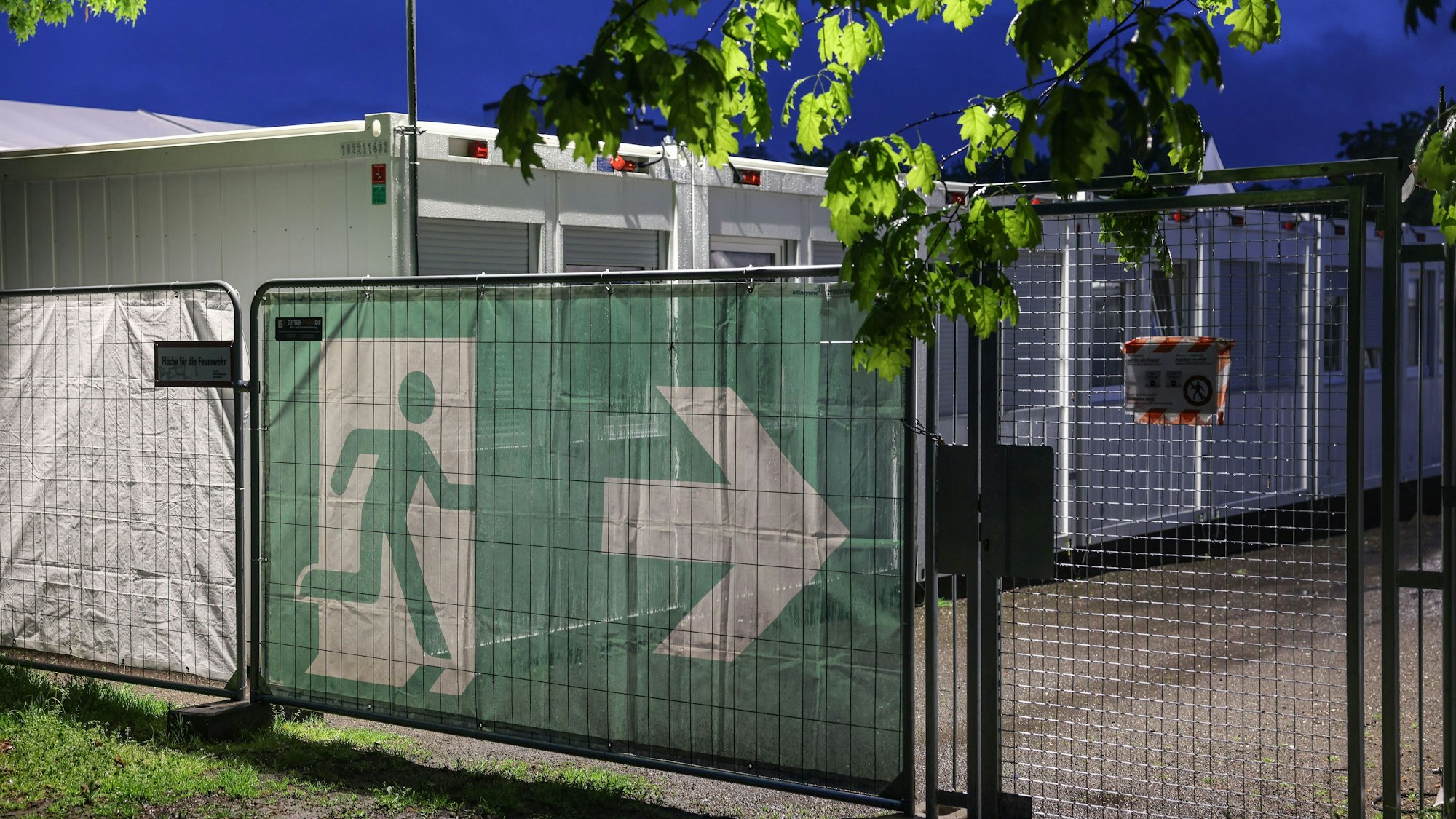 10.05.2023, Nordrhein-Westfalen, Köln: Ein Transparent mit dem Symbol für den Fluchtweg hängt vor Wohncontainer der kommunalen Erstaufnahmeeinrichtung in Köln-Zollstock. Bund und Länder suchen bei einem Spitzentreffen in Berlin nach Lösungen im Streit über die Aufteilung der Kosten für die Versorgung von Flüchtlingen. (Zu dpa: «Gipfel berät über Flüchtlingsfrage») Foto: Oliver Berg/dpa +++ dpa-Bildfunk +++
