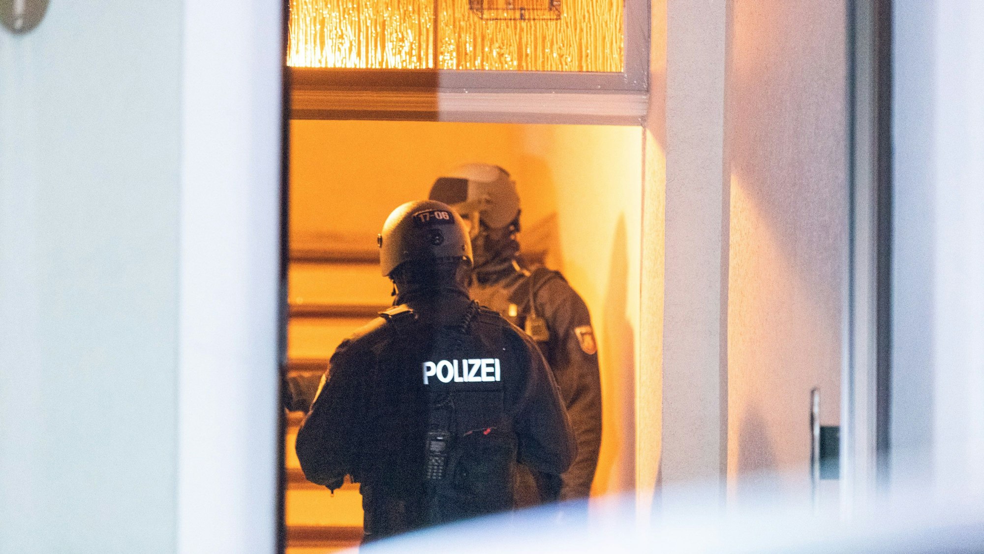 Zwei Polizisten stehen hinter einer Haustür im Treppenhaus.