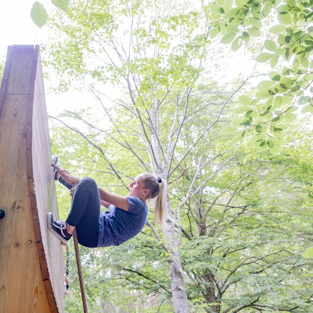 eine Frau klettert in einem Wald eine Wand aus Holz an einem Seil hoch