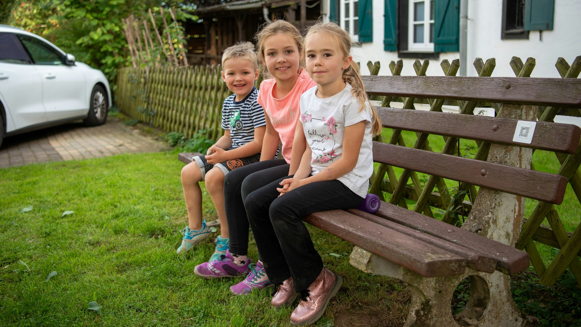 Drei Kinder sitzen auf einer Bank. Im Hintergrund sind ein Haus und ein Auto zu erkennen.