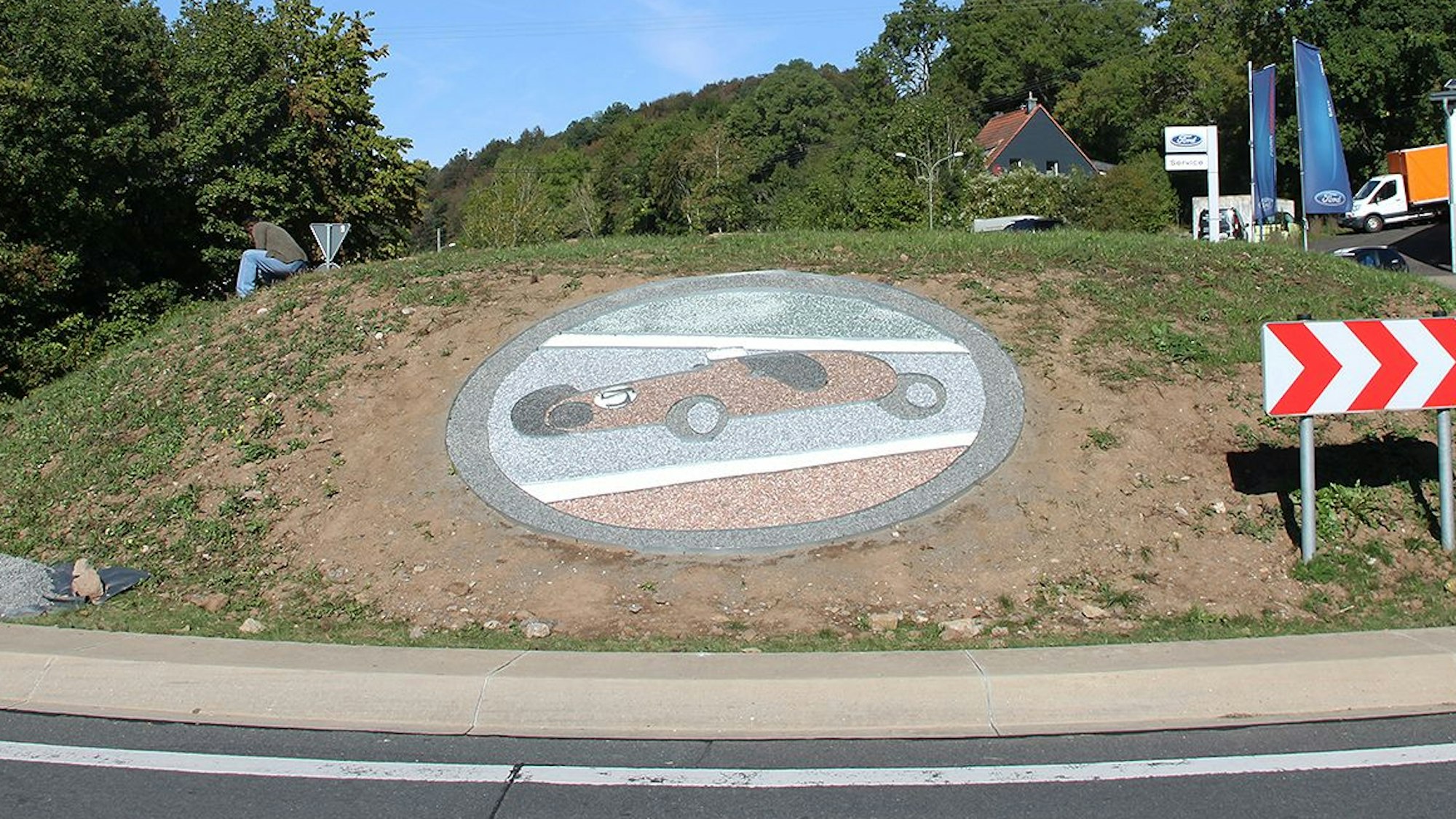 Ein Rennwagen-Motiv aus farbigem Splitt ist an der Abbiegung Richtung Nürburgring auf der Insel des Kreisverkehrs in Ahrhütte zu sehen.