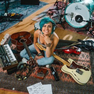 Die Musikerin Charly Klauser sitzt mit Kopfhörern auf einem Teppich, um sie herum sind Rockmusikinstrumente drapiert.