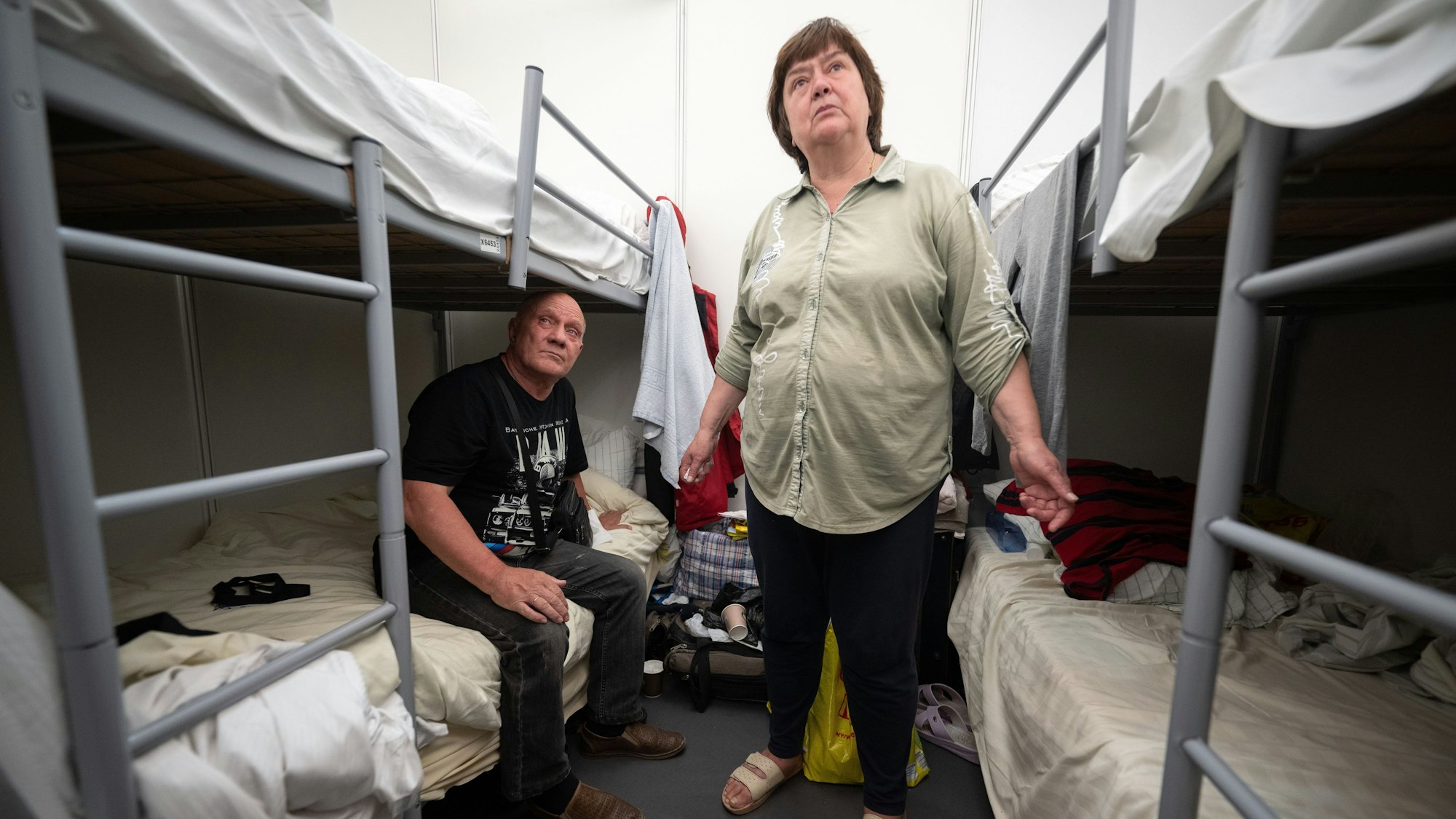 Halina und Oleg aus der Ukraine sitzen und stehen in der Notunterkunft für Geflüchtete am ehemaligen Flughafen Tegel zwischen ihren Betten in einem 12-Bett-Zimmer.