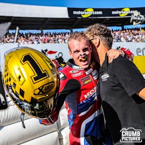 Florian Alt jubelt und umarmt einen Teamkollegen. In der Hand hält er einen goldenen Motorradhelm.