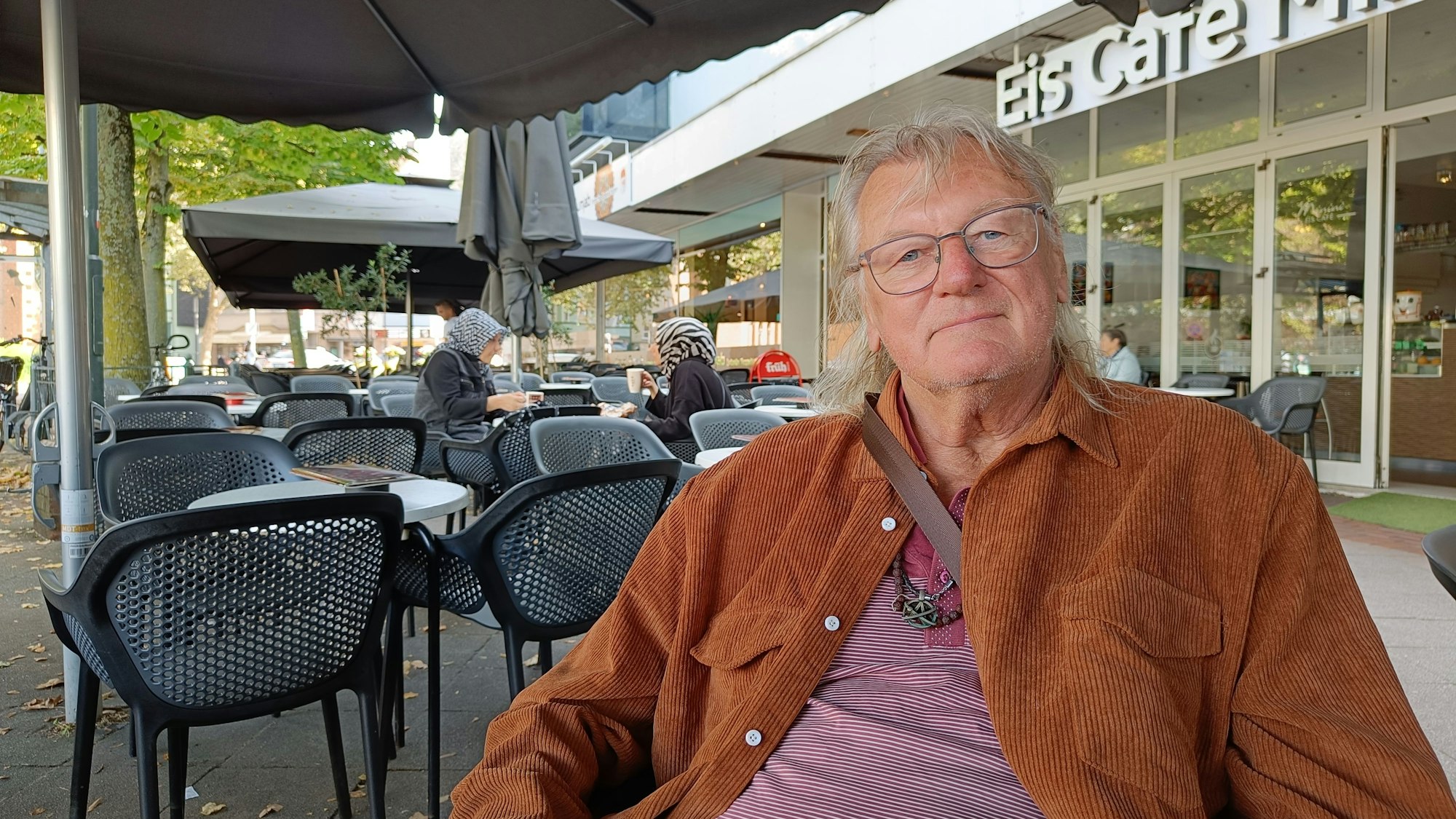 Ein Mann mit brauner Jacke und grauen Haaren sitzt vor einem Eiscafé.