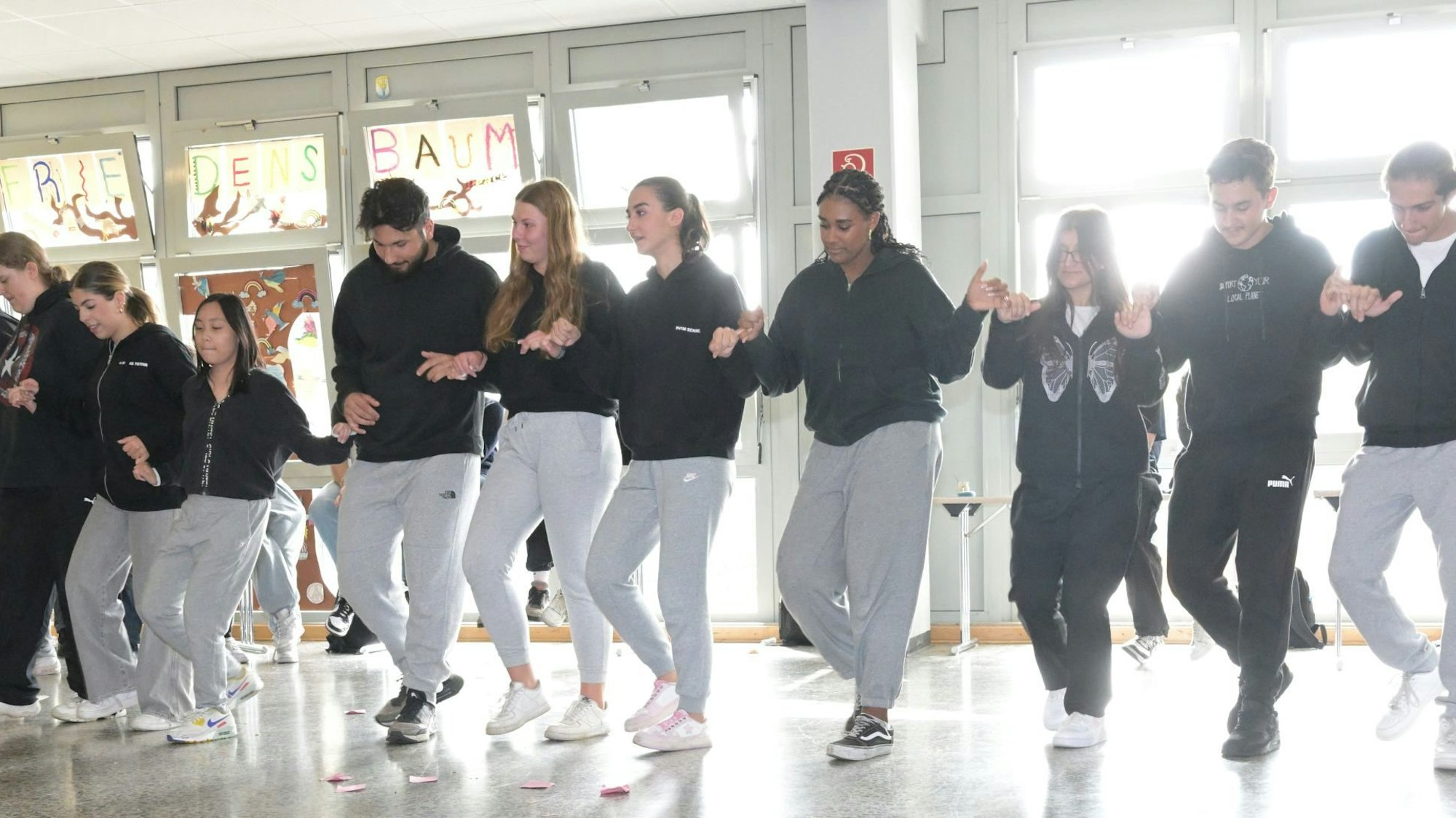 Schülerinnen und Schüler, alle mit einem schwarzen Sweatshirt und einer Sporthose angezogen, halten sich an den Händen und machen dabei Tanzschritte. 