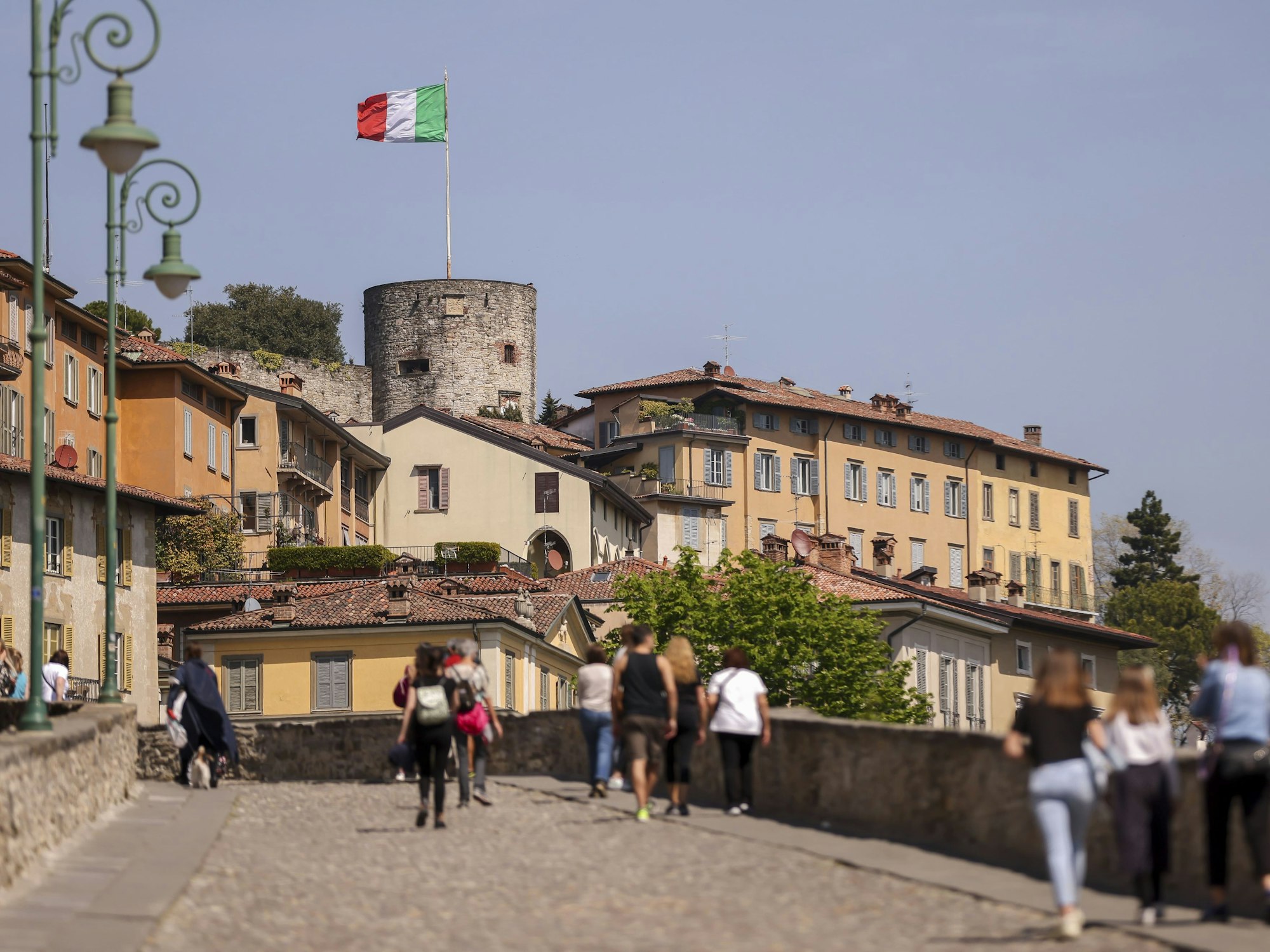 Touristen und Touristinnen gehen über eine Brücke hinauf zur Altstadt (Citta Alta) auf den Hügel, hier im April 2022 in Bergamo (Italien).