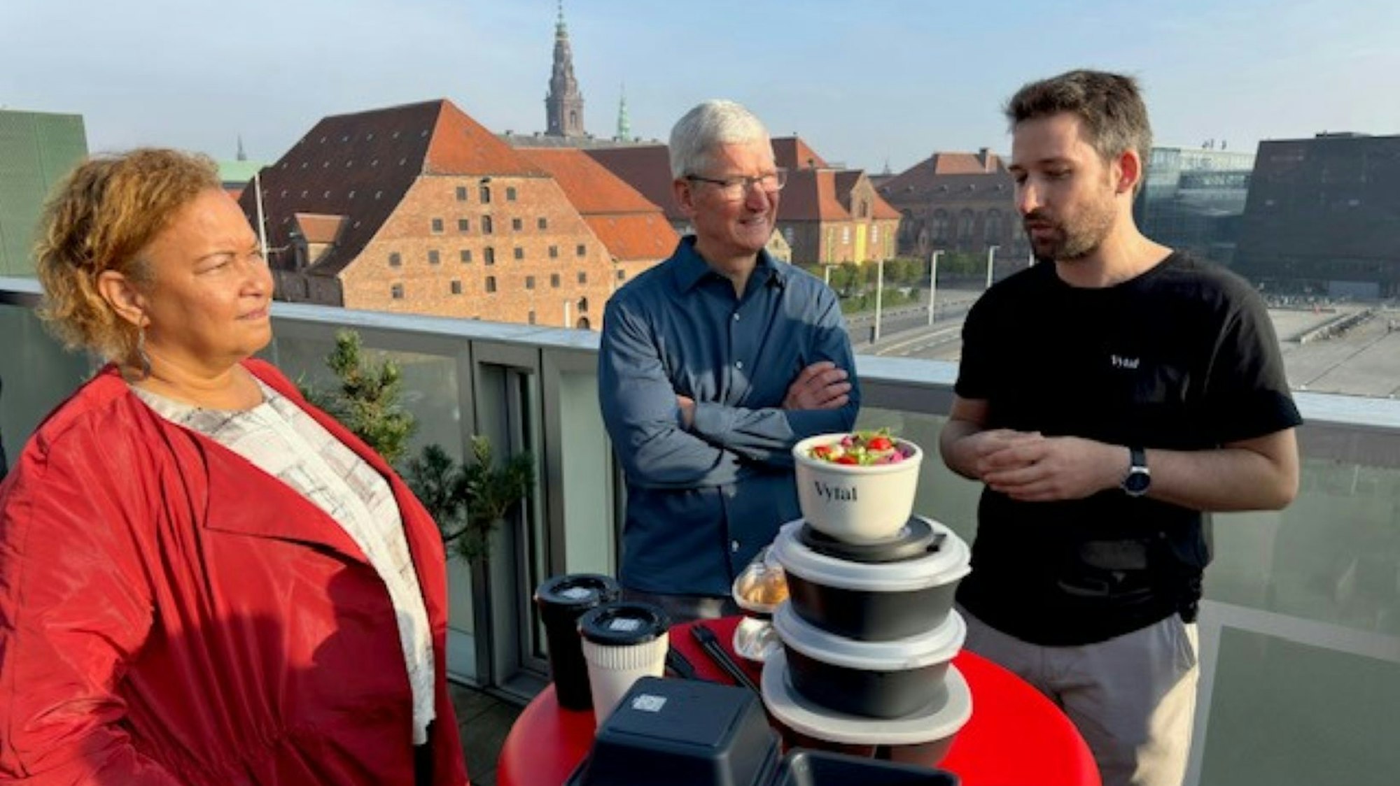 Apple-Chef Tim Cook, der Kölner Tim Breker und Lisa P. Jackson, ebenfalls von Apple stehen auf einer Dachterrasse an einem runden Tisch, auf dem Mehrwegschalen gestapelt sind.