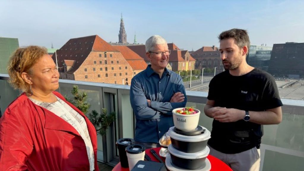 Apple-Chef Tim Cook, der Kölner Tim Breker und Lisa P. Jackson, ebenfalls von Apple stehen auf einer Dachterrasse an einem runden Tisch, auf dem Mehrwegschalen gestapelt sind.