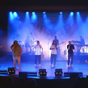 Eine Band mit vier Sängern im Vordergrund und mehreren Instrumentalisten steht auf der blau illuminierten Bühne des Euskirchener Theaters.