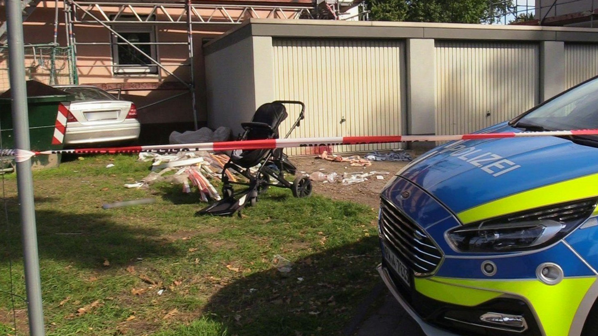 Ein beschädigter Kinderwagen steht auf einem Rasen neben mehreren Garagen, dahinter, an einer Hauswand, steht ein Auto. Die Polizei hat den Bereich mit Flatterband abgesperrt.