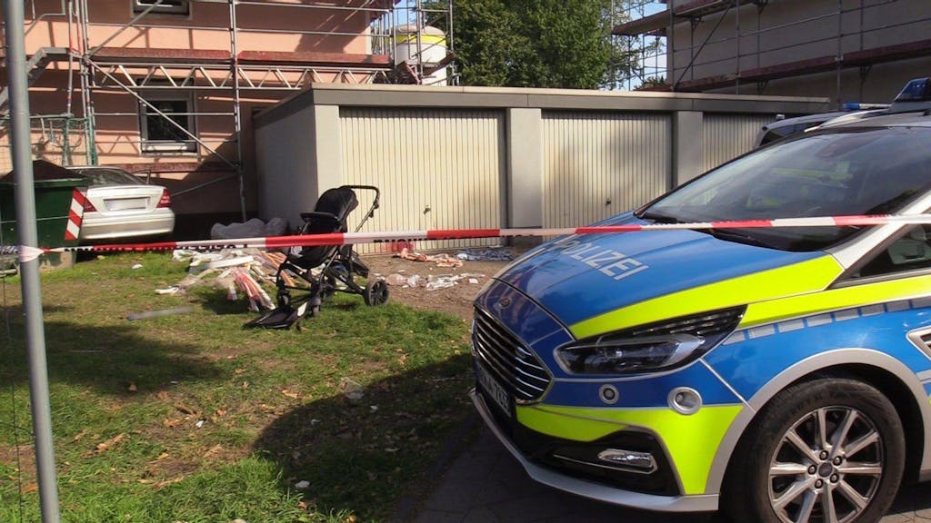 Ein beschädigter Kinderwagen steht auf einem Rasen neben mehreren Garagen, dahinter, an einer Hauswand, steht ein Auto. Die Polizei hat den Bereich mit Flatterband abgesperrt.&nbsp;