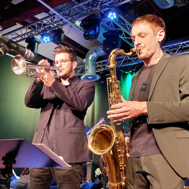 Ein junger Mann mit Trompete und ein weiterer mit Saxofon stehen im Rampenlicht auf einer Bühne, beide tragen Anzüge.