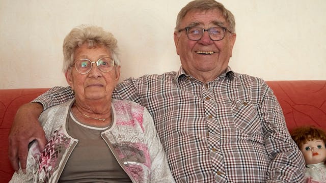 Ein Ehepaar sitzt auf einem roten Sofa: Fritz Bahne legt einen Arm um seine Ehefrau Angelika, mit der er seit 65 Jahren verheiratet ist.