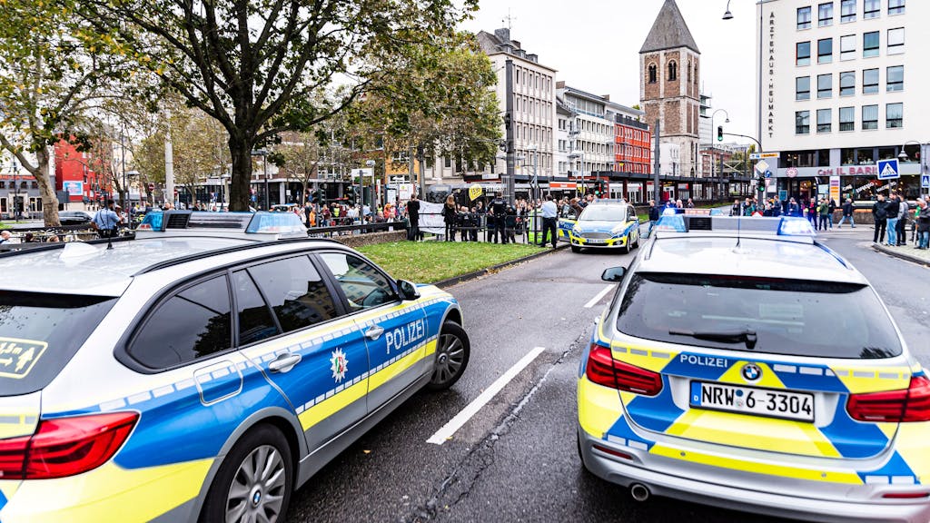 Zwei Polizei-Autos stehen am Kölner Heumarkt, eine Demo im Hintergrund wird überwacht.