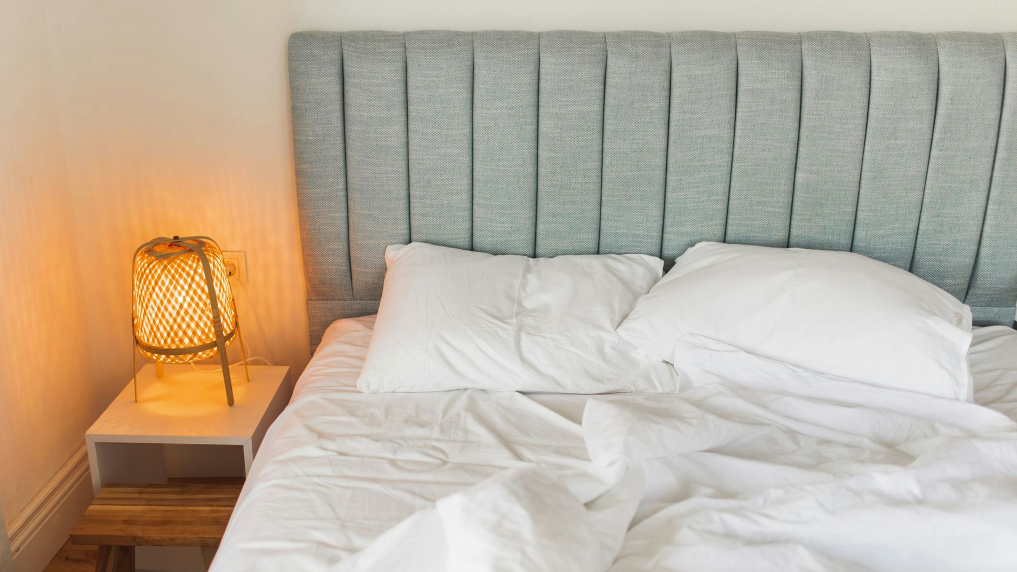 Auf dem Foto sieht man ein graues Bett, links daneben steht ein Nachtschrank mit einer eingeschalteten Lampe darauf.
