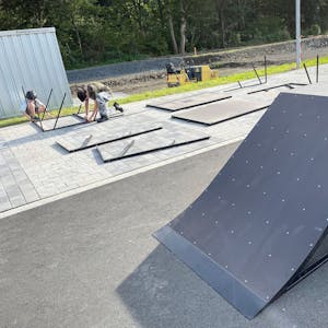 Die Rampe eines mobilen Skateparks ist zu sehen. Im Hintergrund schrauben zwei Jugendliche weitere Elemente zusammen.&nbsp;