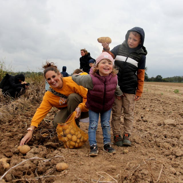 Eine Mutter kniet auf einem Acker und sammelt Kartoffeln ein. Ihre beiden Kindern grinsen in die Kamera.
