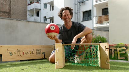 Ein Mann hält einen Ball in den Händen und kniet vor einem kleinen Fußballtor aus Holz.
