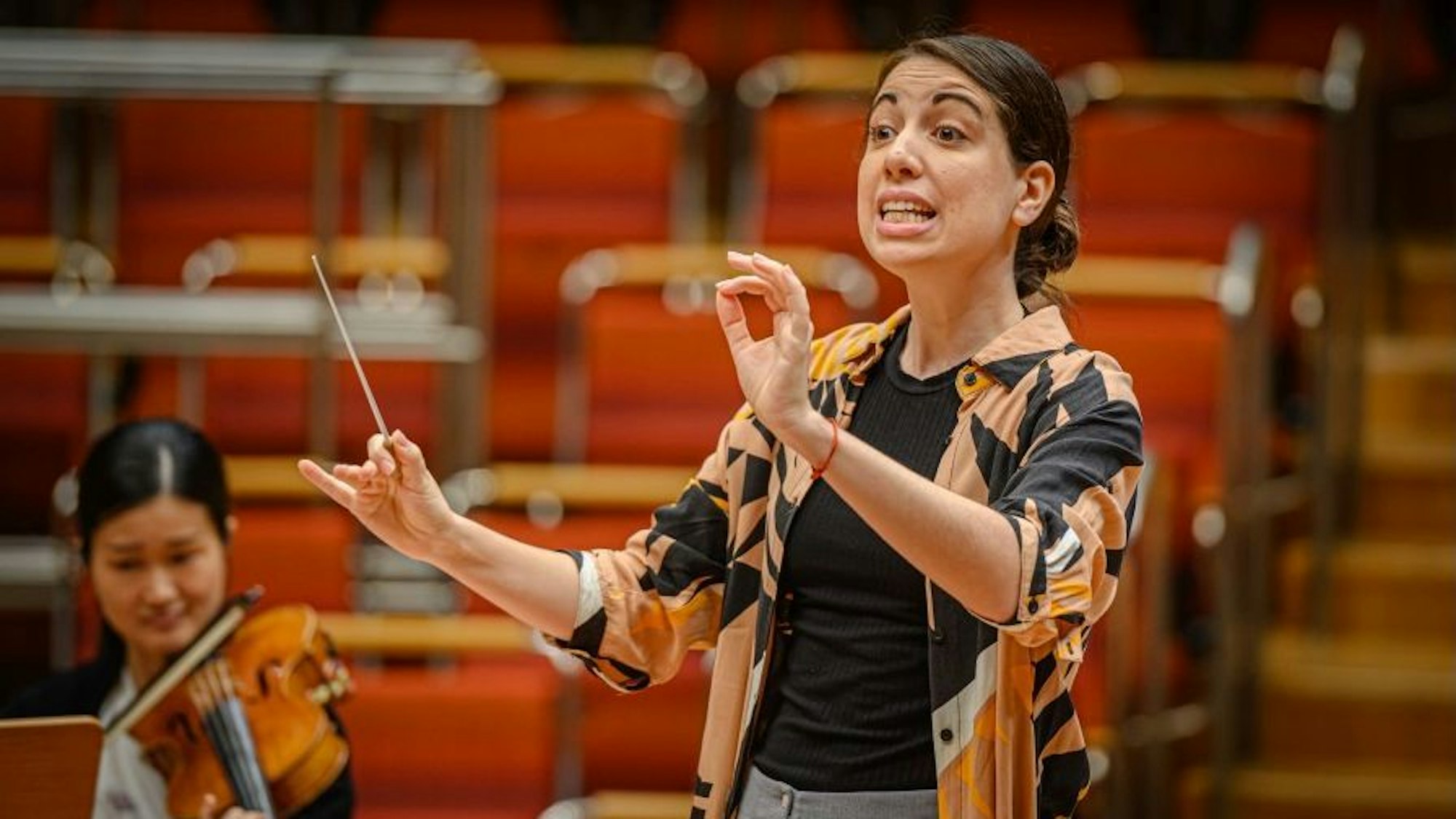 Die Dirigentin Anna Castro Grinstein in der Kölner Philharmonie. Sie schaut auf ihr Orchester und hat die Hände zum Dirigat erhoben, in der rechten Hand hält sie einen Taktstock.